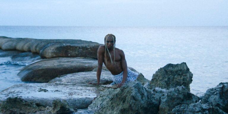 A film still showing a person sitting on rocks in a body of water.