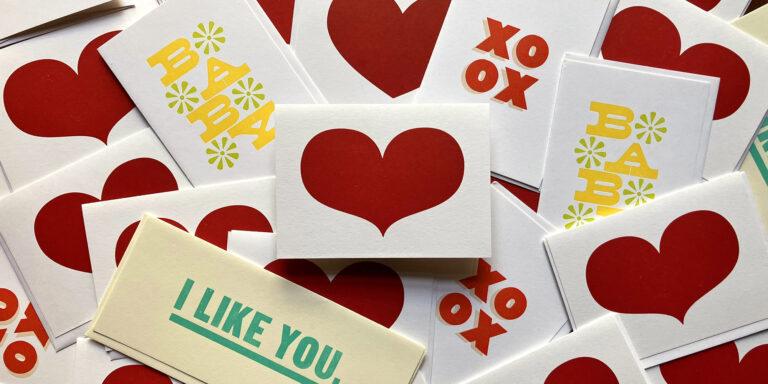A photograph of various greeting cards that feature red hearts, the word “Baby” in yellow, the letters “XOXO” in red, and “I Like You” in green