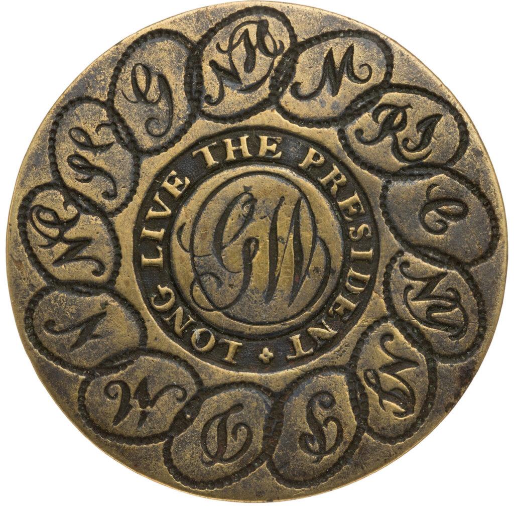 A round brass button featuring the initials &ldquo;GW&rdquo; in the center (for George Washington), surrounded by the words &ldquo;Long Live the President&rdquo;. Around the edge of the button are the initials of each of the 13 original colonies (states) linked together in circles.